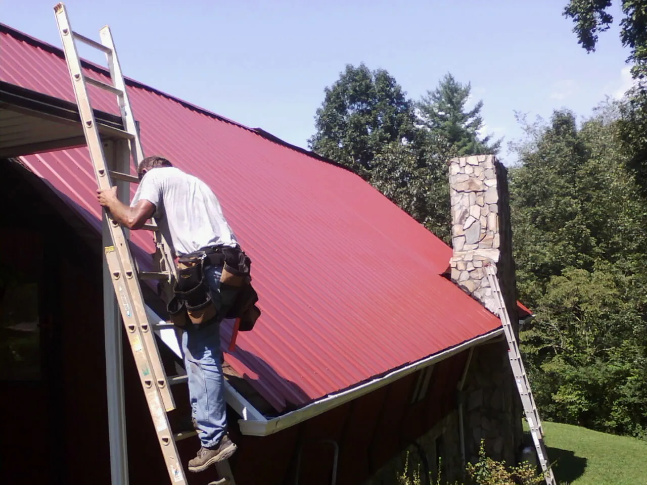 Roofer on a ladder beside a red metal roof and stone chimney — Soffit & Fascia Repair by Roofing Brothers in Springwater